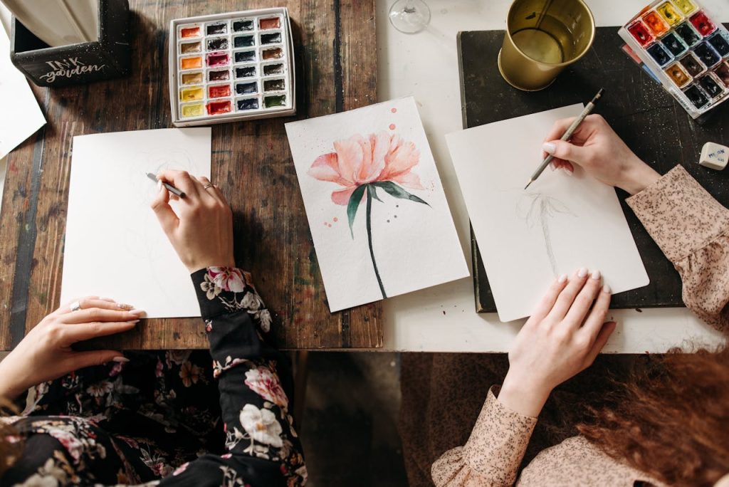 women-drawing-on-white-paper-6925172 Overhead view of artists drawing floral designs with watercolors and pencils, indoors.